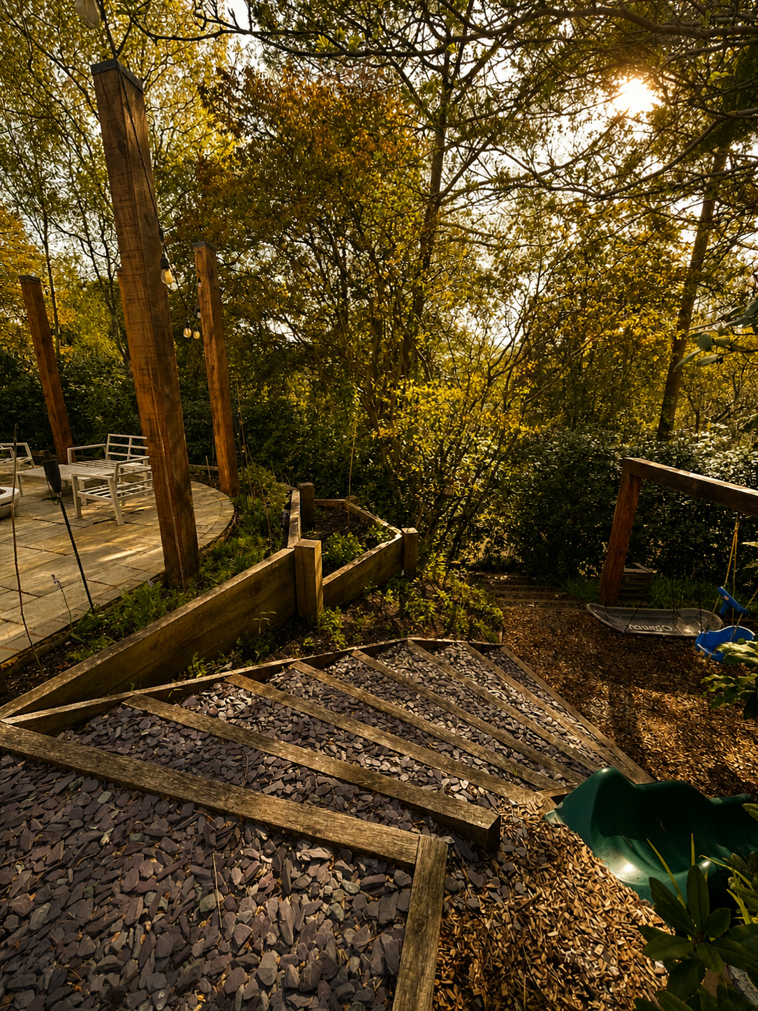 A backyard with a stone patio area, wooden stairs leading down to a gravel path, surrounded by trees and foliage, with outdoor seating including white chairs, and a swing on a bamboo frame, with sunlight filtering through the trees.