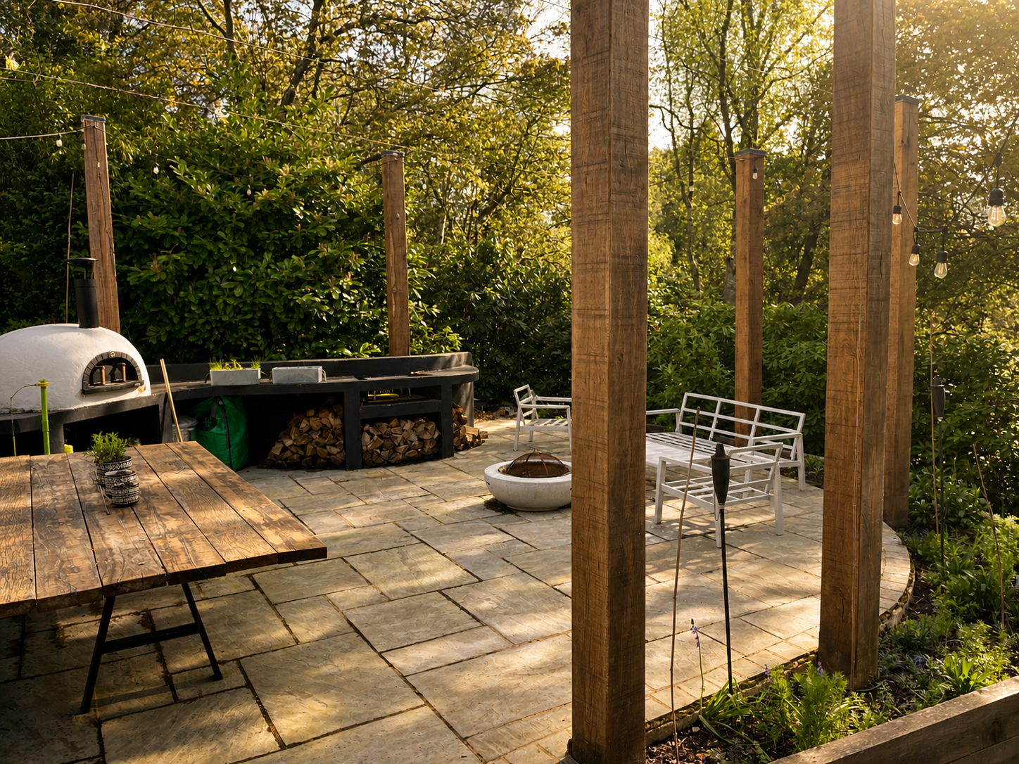 A backyard patio with a wood table, two white benches, a stone fire bowl, a wood-fired oven, and a wood storage area, surrounded by lush green trees and shrubbery.
