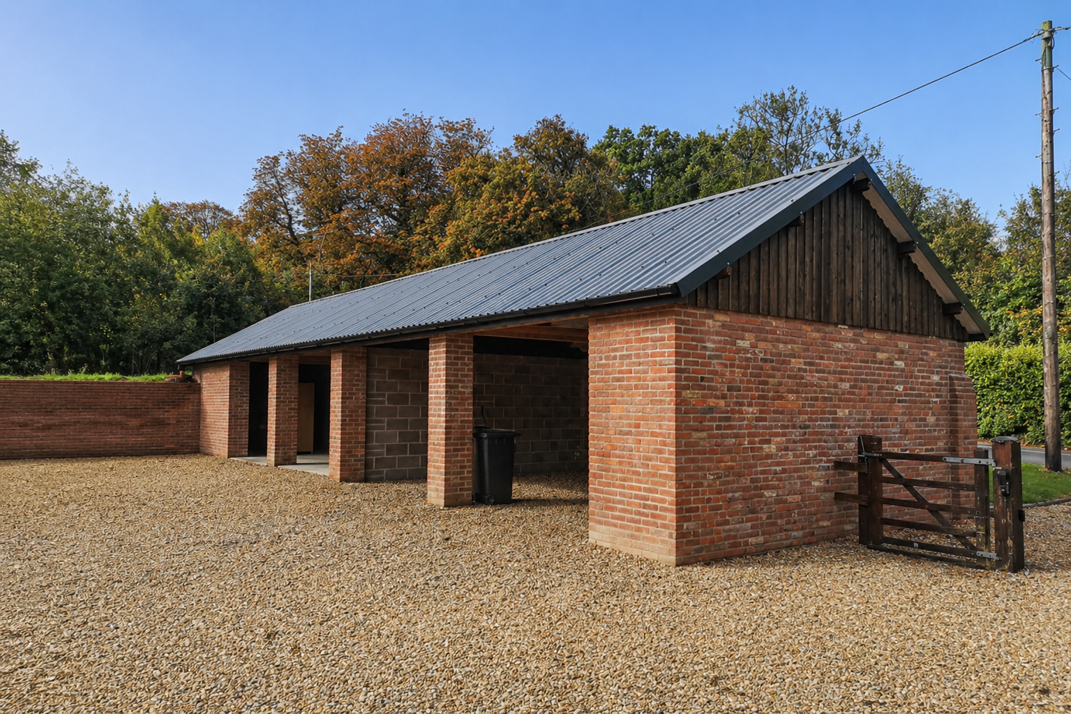 A brick building with a metal roof on a gravel yard, surrounded by trees with autumn foliage, and a wooden gate on the right side.