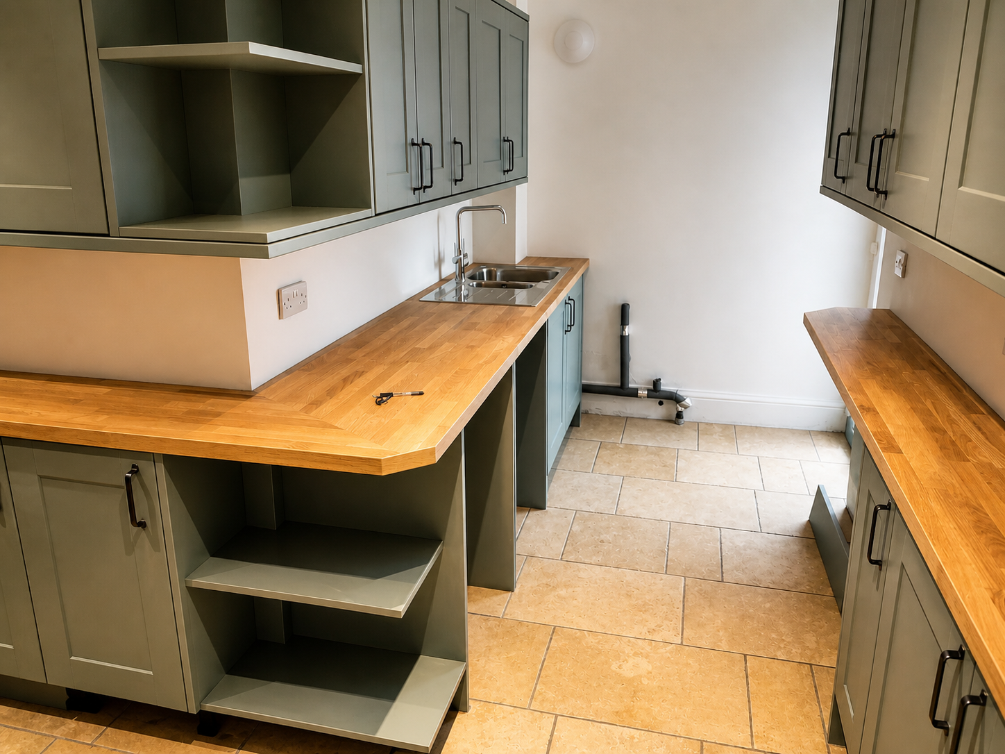 Empty kitchen with green cabinets, wooden countertops, stainless steel sink, tiled floor, and an exposed pipe in the corner.