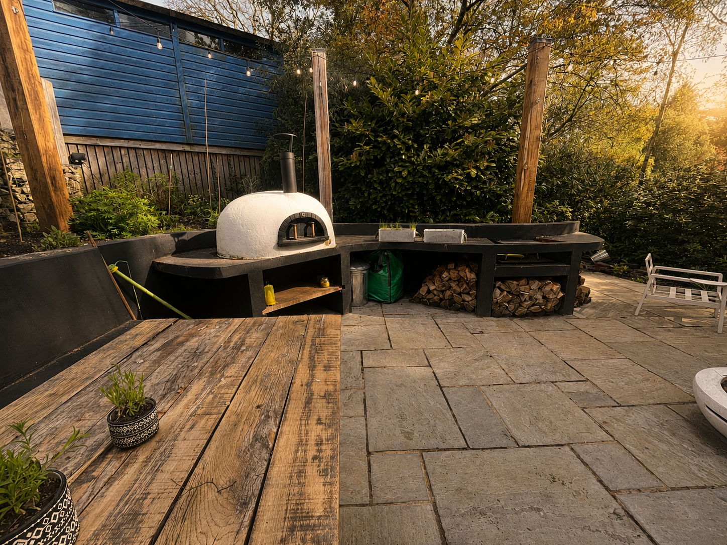Outdoor kitchen with pizza oven, wood pile, and seating area on stone patio with trees and fence in background at sunset.
