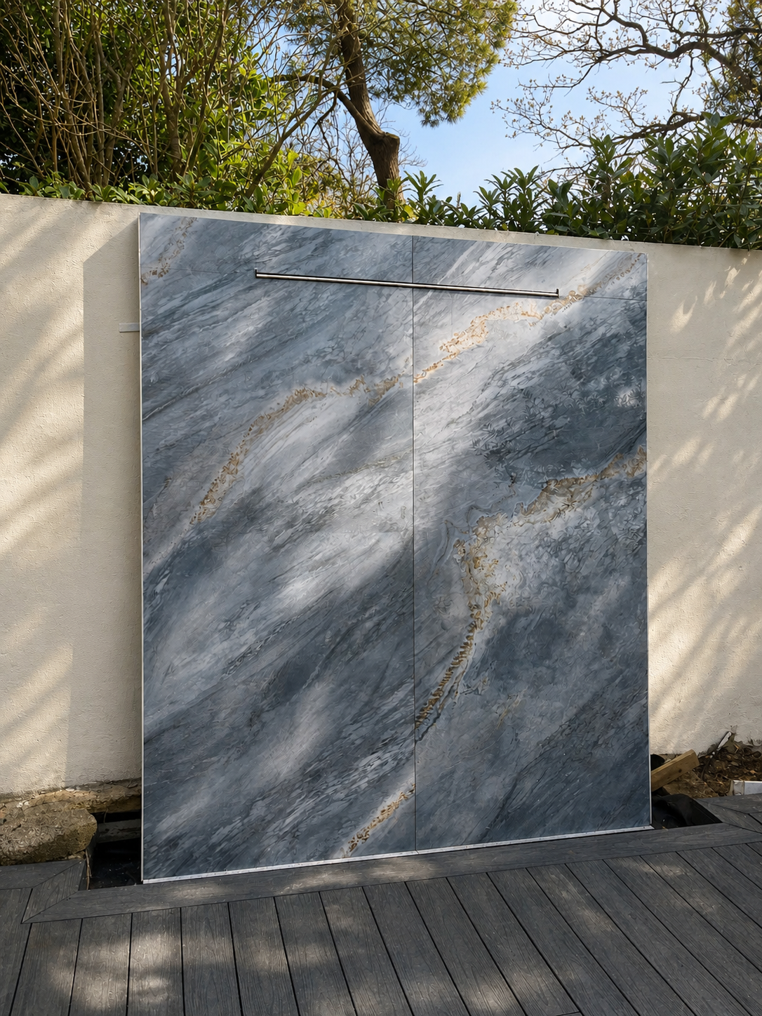 An outdoor shower with a marble wall, mounted on a beige section of wall, surrounded by trees and shadows, with a wooden deck floor.