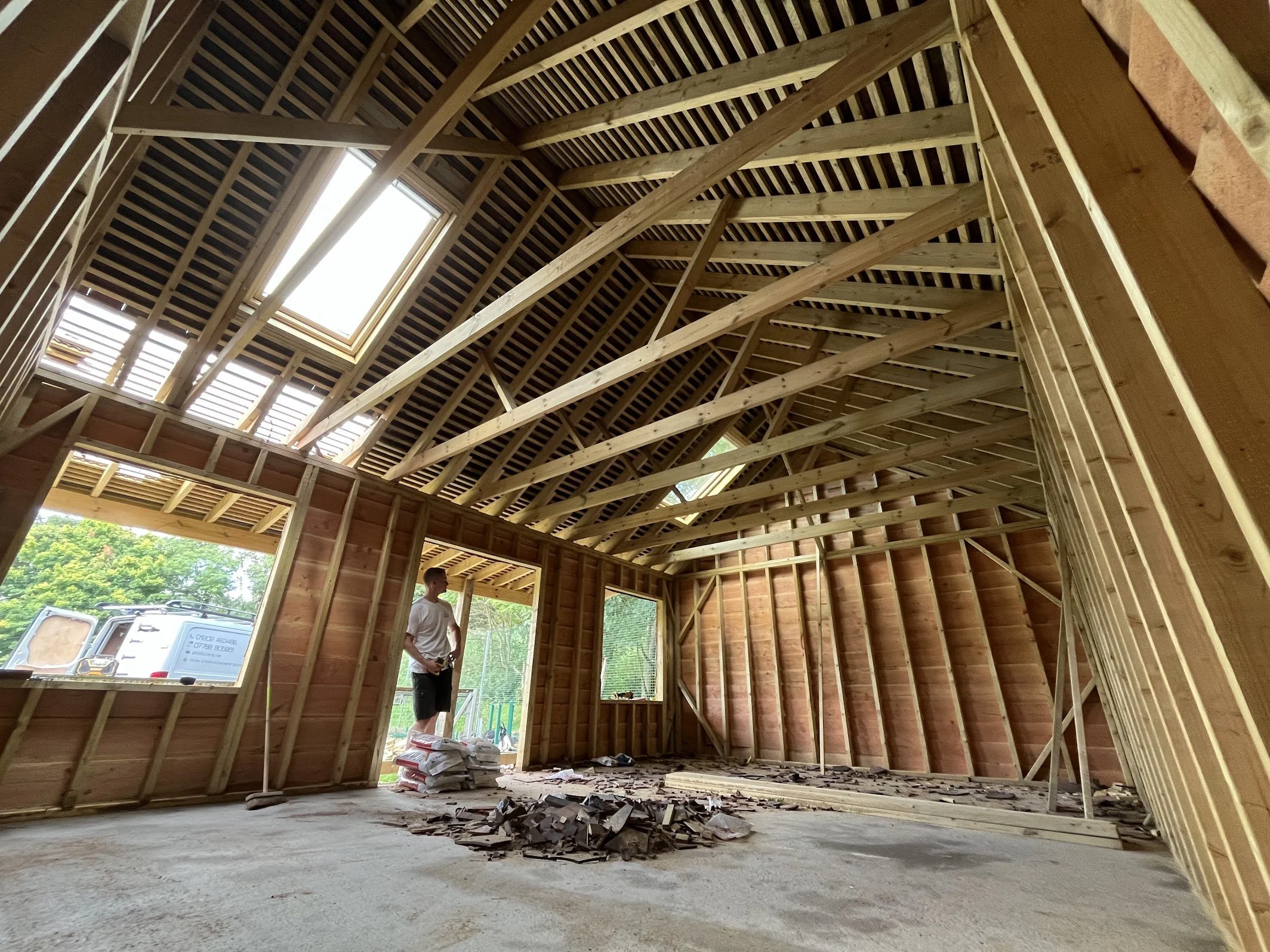 Interior view of a building under construction with exposed wooden framing, a skylight, and a person standing inside observing the construction process.