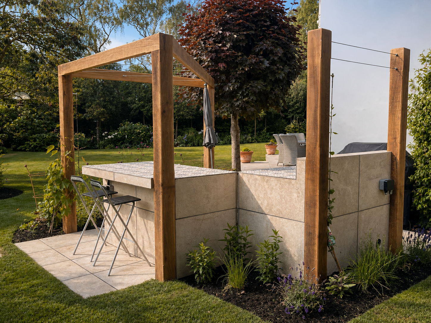 An outdoor backyard kitchen with a tiled counter, wooden pergola framework, and patio furniture, surrounded by greenery and trees.