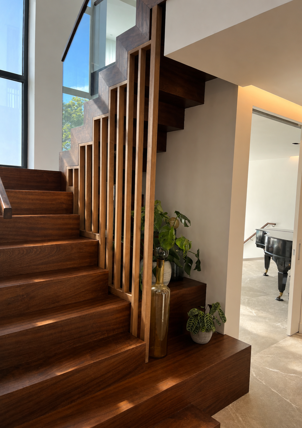 Indoor wooden staircase with vertical slats railing, next to a large window, with potted plants and a doorway leading to a room with a grand piano visible.