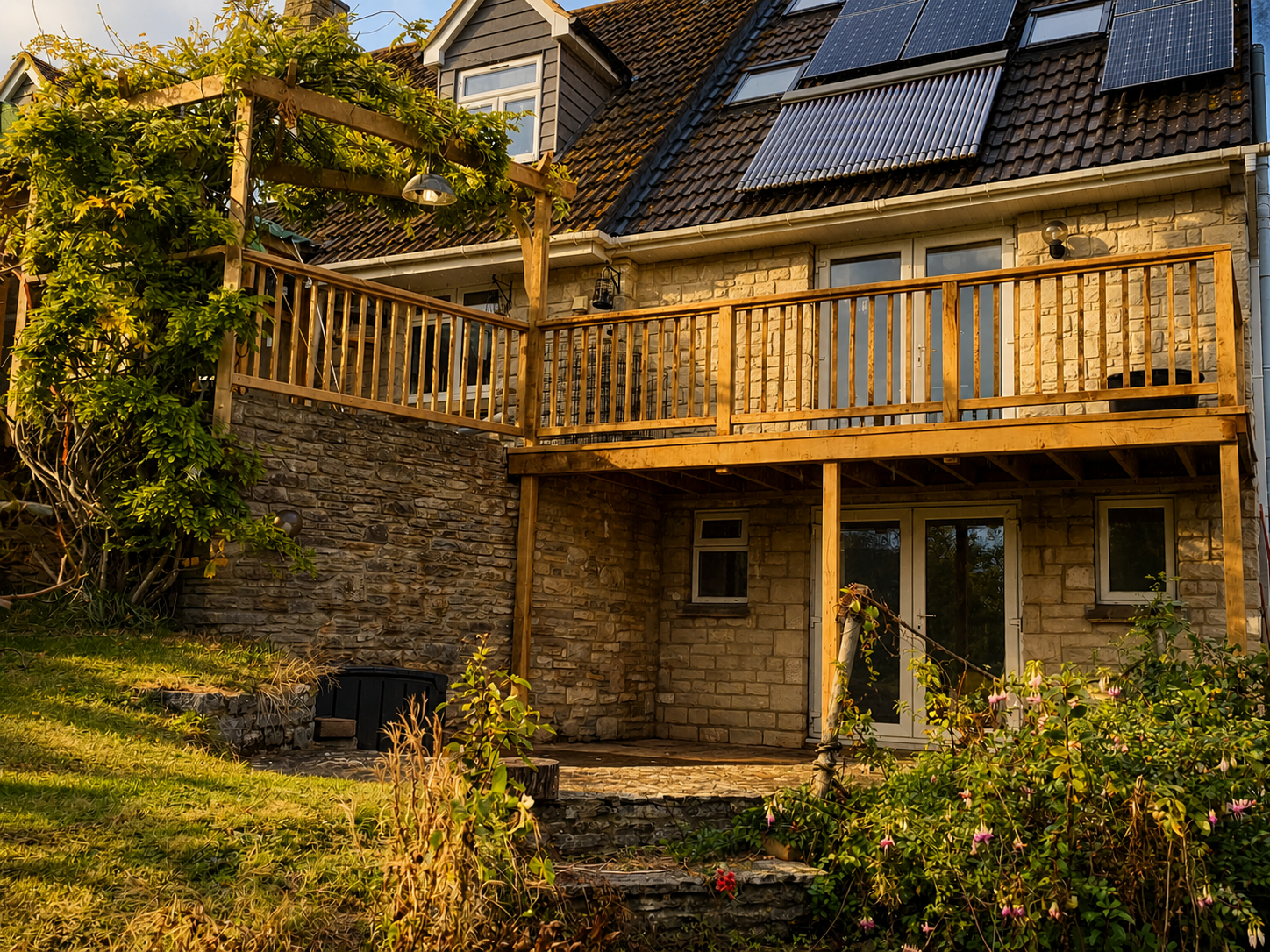 The back of a house with a stone exterior, a wooden balcony, and solar panels on the roof, surrounded by greenery and plants.