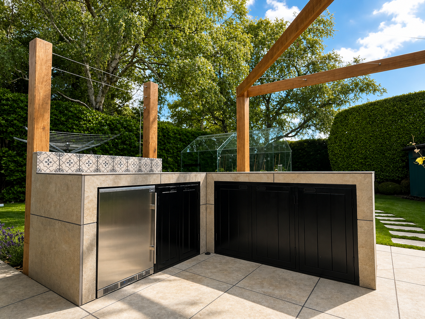 An outdoor kitchen with a tiled counter and black cabinets, a stainless steel mini refrigerator, in a backyard with green trees, grass, and stepping stones, under a blue sky with some clouds.
