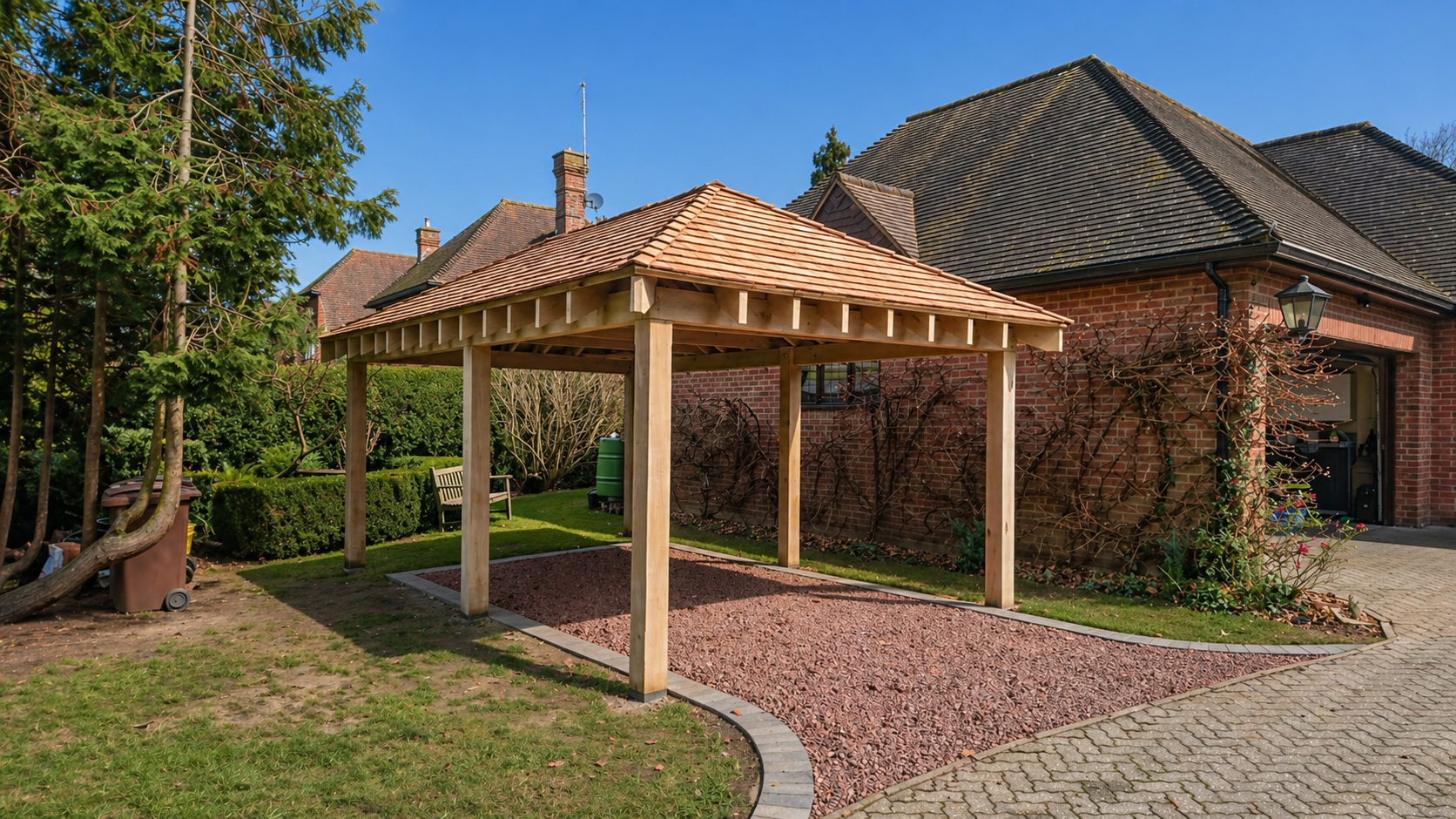 Small wooden gazebo with a shingled roof, located in a backyard with a brick house and garden surrounding it. There is a gravel area and a paved pathway nearby.