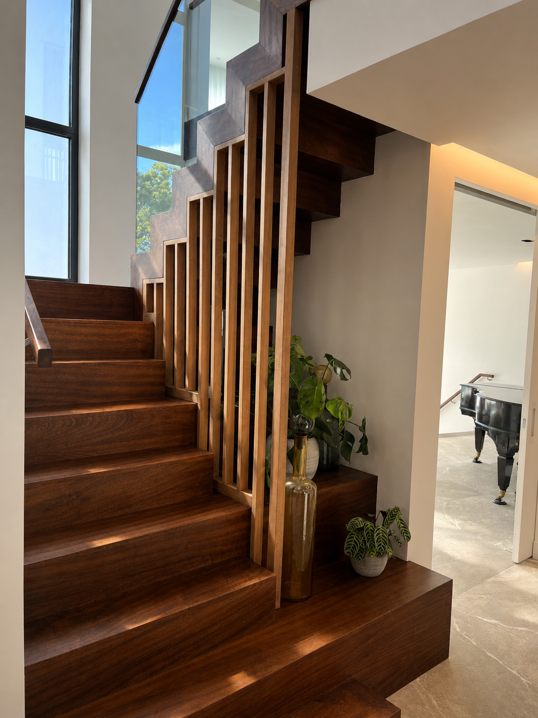 Interior of a modern house featuring a wooden staircase with vertical balustrades, large windows allowing natural light, potted plants, and a glimpse of a black grand piano in another room.