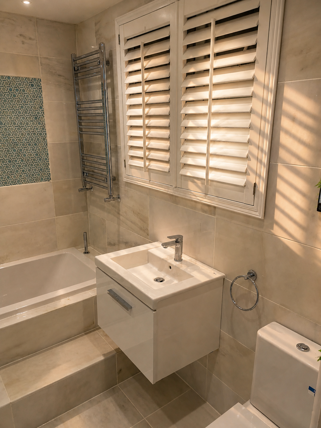 Bathroom with beige tiled walls and floor, a white window with closed blinds, a small white sink with a chrome faucet, a white toilet, and a heated towel rack on the wall.
