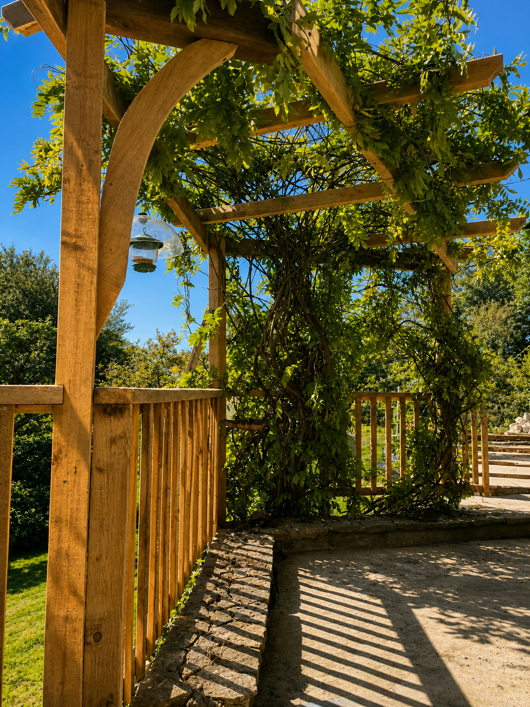 Wooden pergola with green vines and leaves, hanging bird feeder, and a wooden railing on a sunny day with blue sky.