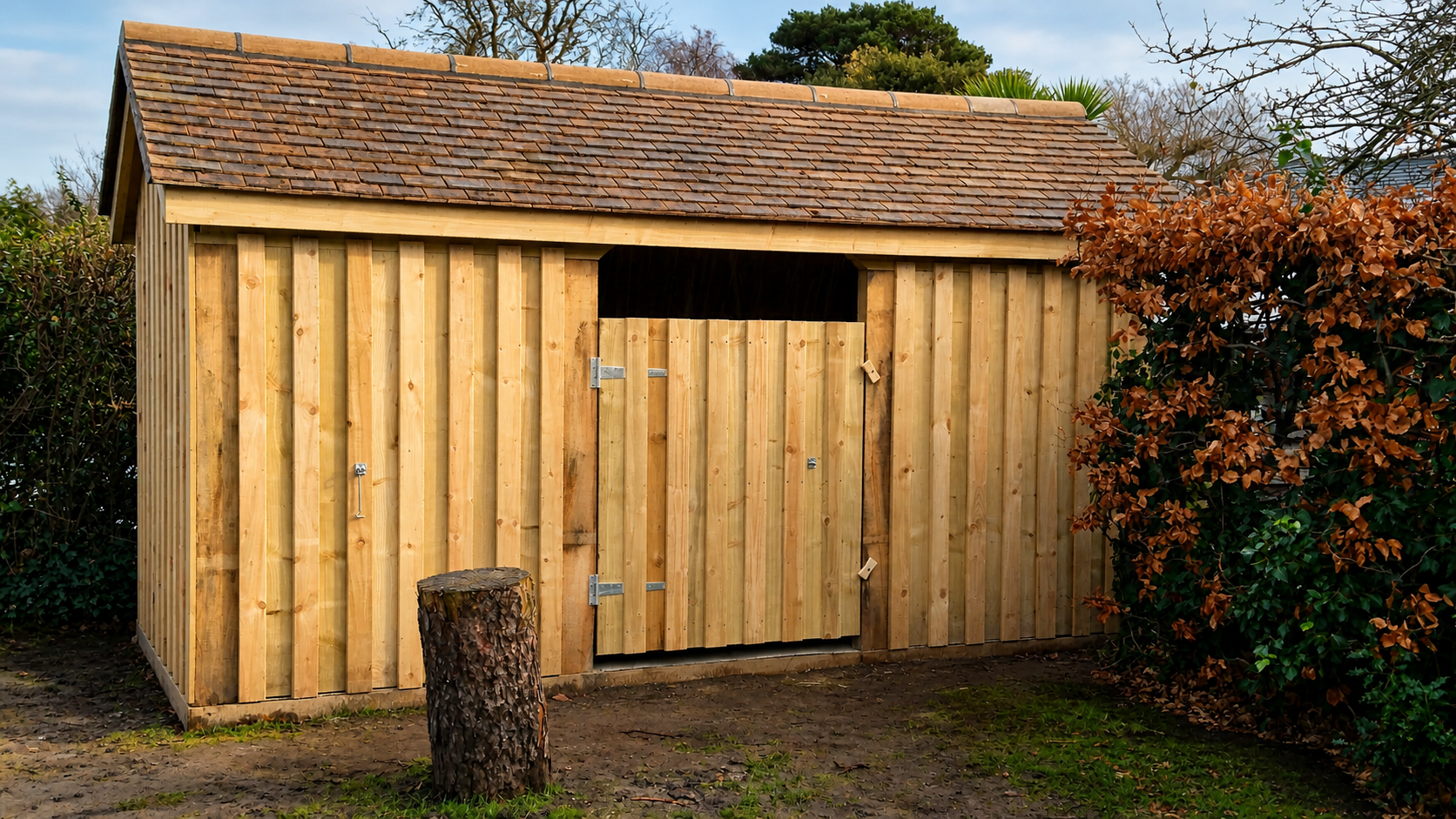 New wooden garden shed with a shingled roof, partially open door, surrounded by brown and green bushes, a small tree trunk in front, and a muddy ground.