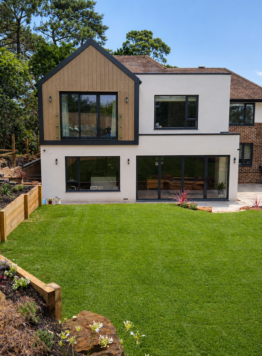 A modern two-story house with a white exterior, black window frames, a brown wooden section on the upper level with a large window and sliding door, a well-maintained green lawn, flower beds with colorful plants and gardening tools, and tall trees in the background under a partly cloudy blue sky.