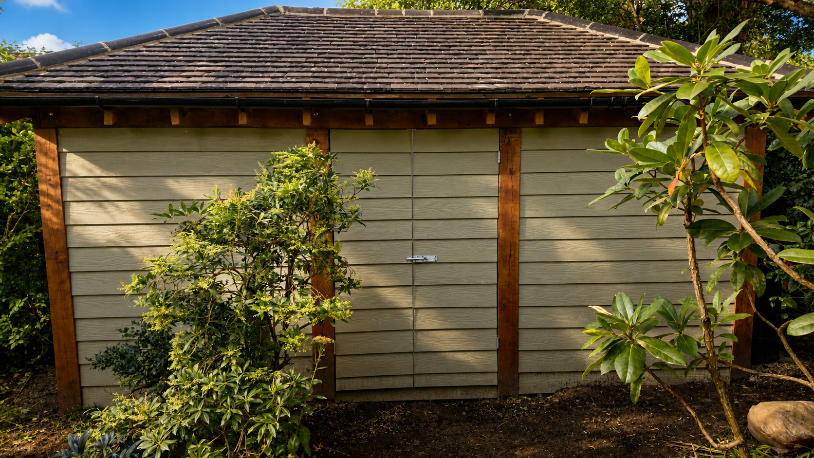 A small wooden shed with green siding and a shingled roof, surrounded by bushes and plants, under a partly sunny sky.
