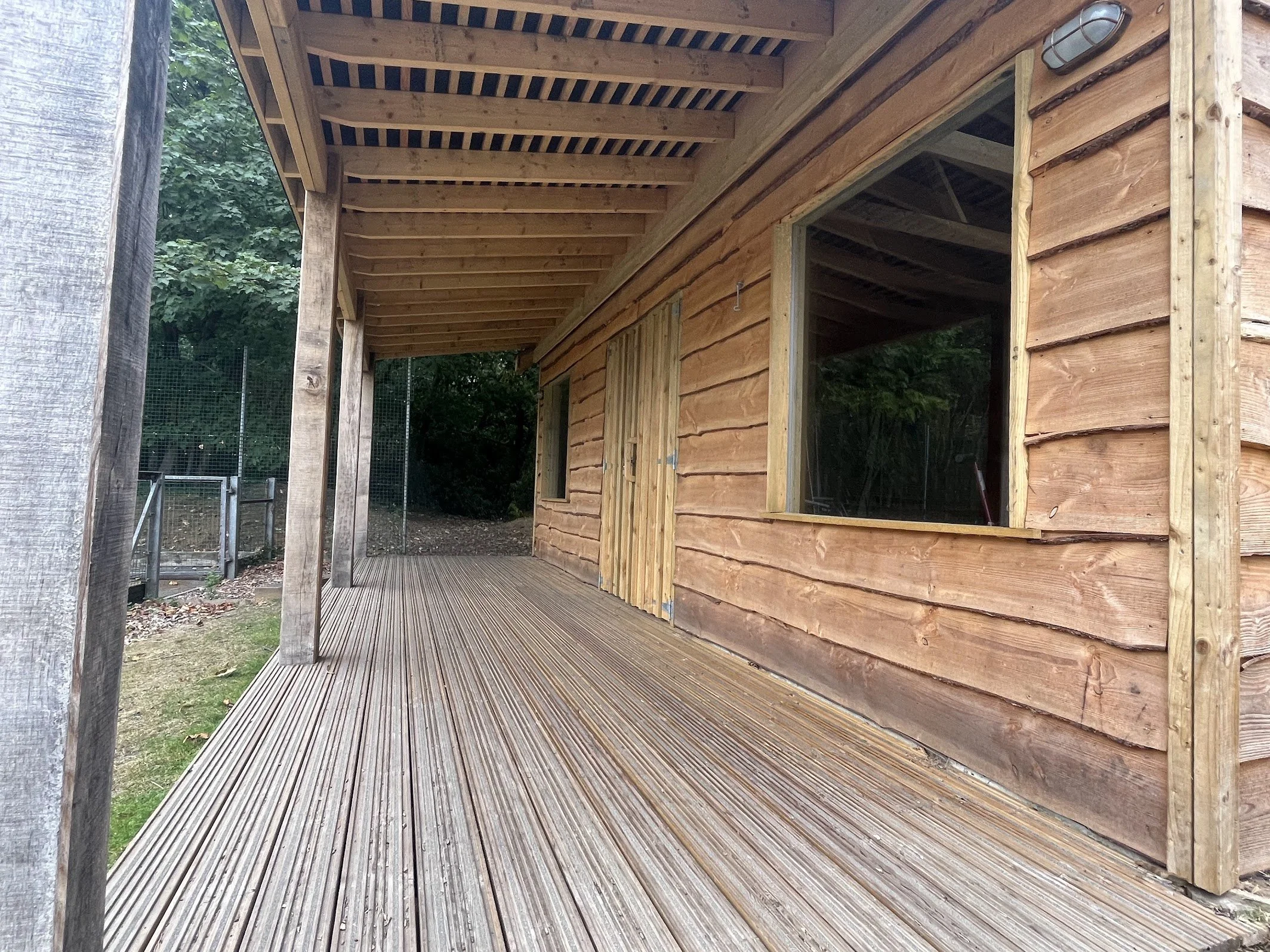 View of a wooden house exterior with large windows, a wooden deck, and a partially built overhang or porch area surrounded by a green, wooded area with some fencing in the background.