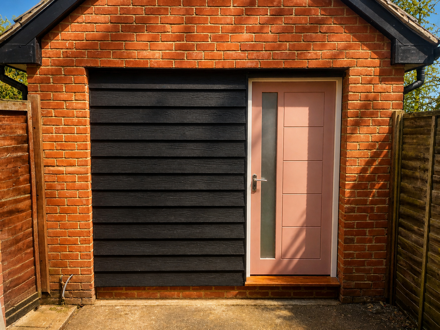 Exterior view of a small brick building with a pink door and black siding, surrounded by wooden fencing.