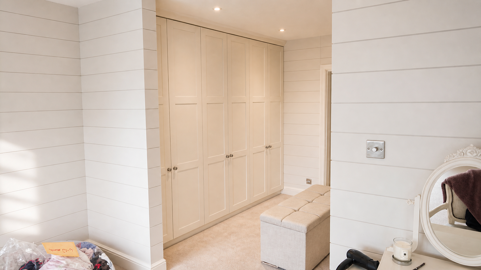 A bedroom corner with white shiplap walls, built-in white closet cabinets, a beige tufted ottoman, and a dressing table with a mirror, candle, and hairdryer.