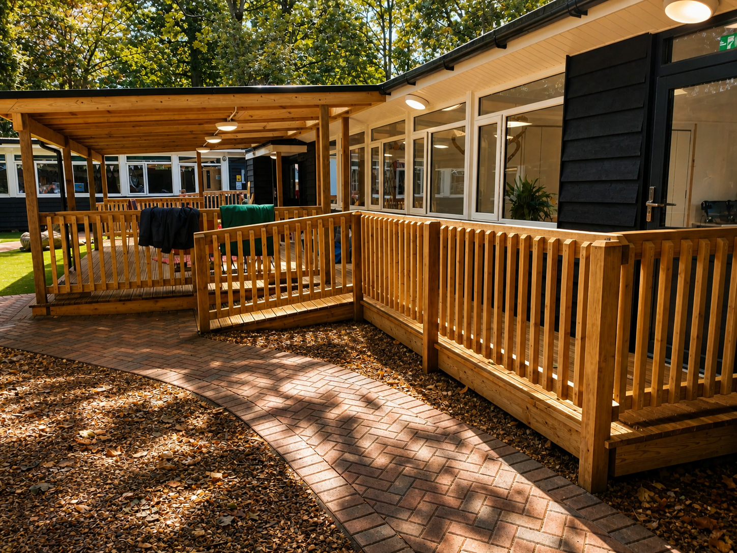 Wooden porch with railing and steps, attached to a building with large windows, surrounded by trees and a brick pathway with fallen leaves.