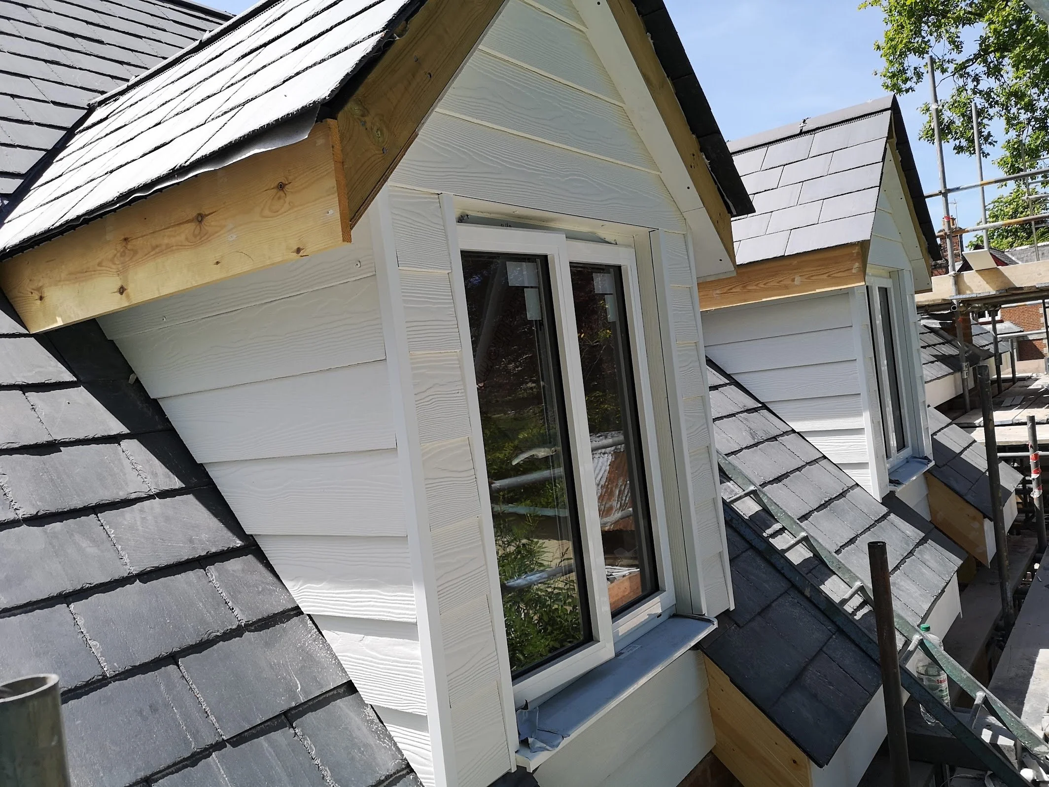 The image shows the exterior of a house under renovation with newly installed white siding and a steep gray tiled roof. Two small dormer windows are visible, with one of them surrounded by scaffolding and construction materials.