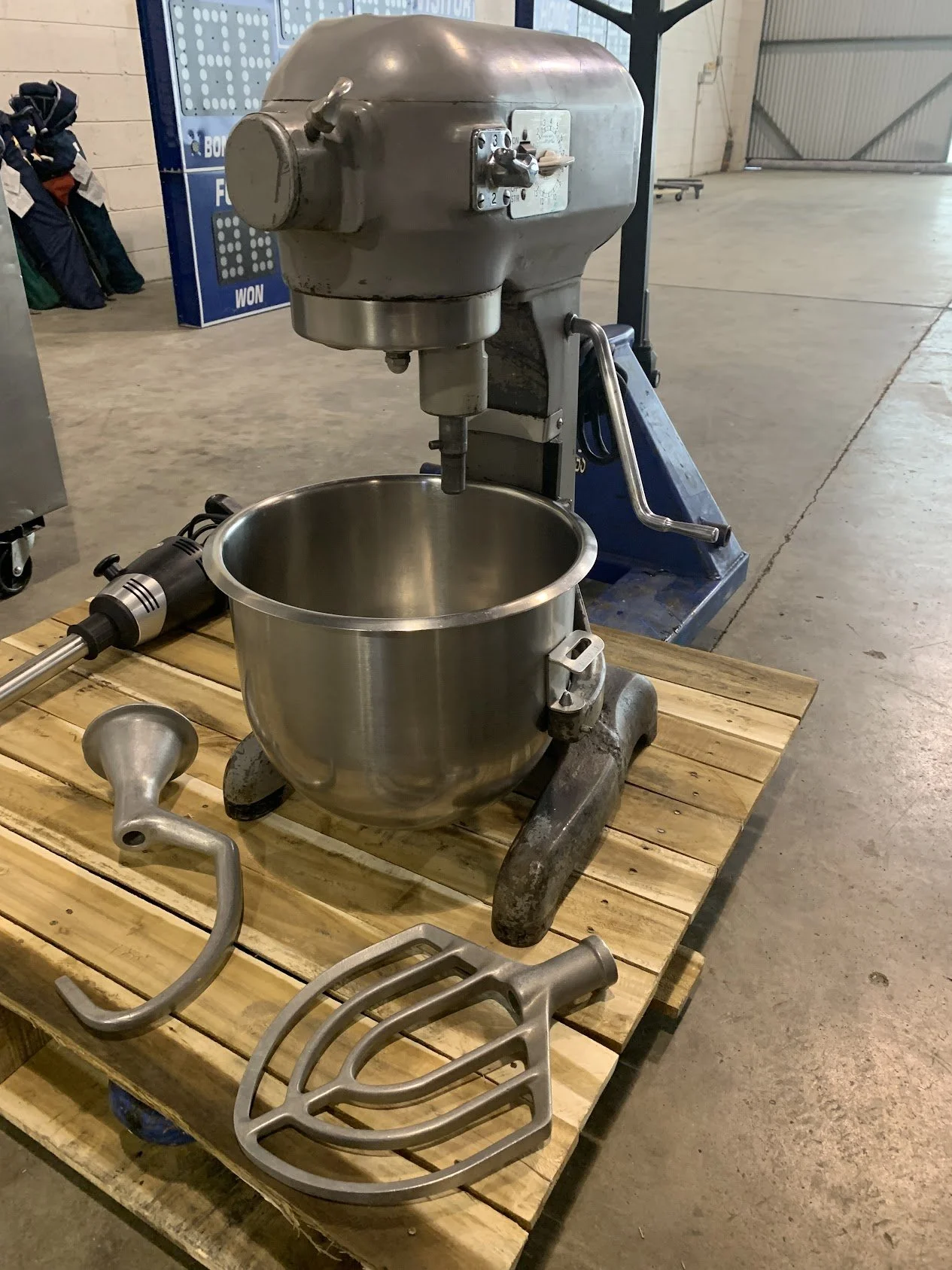 A vintage metal stand mixer on a wooden pallet, with metal attachments and bowls, in an industrial or warehouse setting.