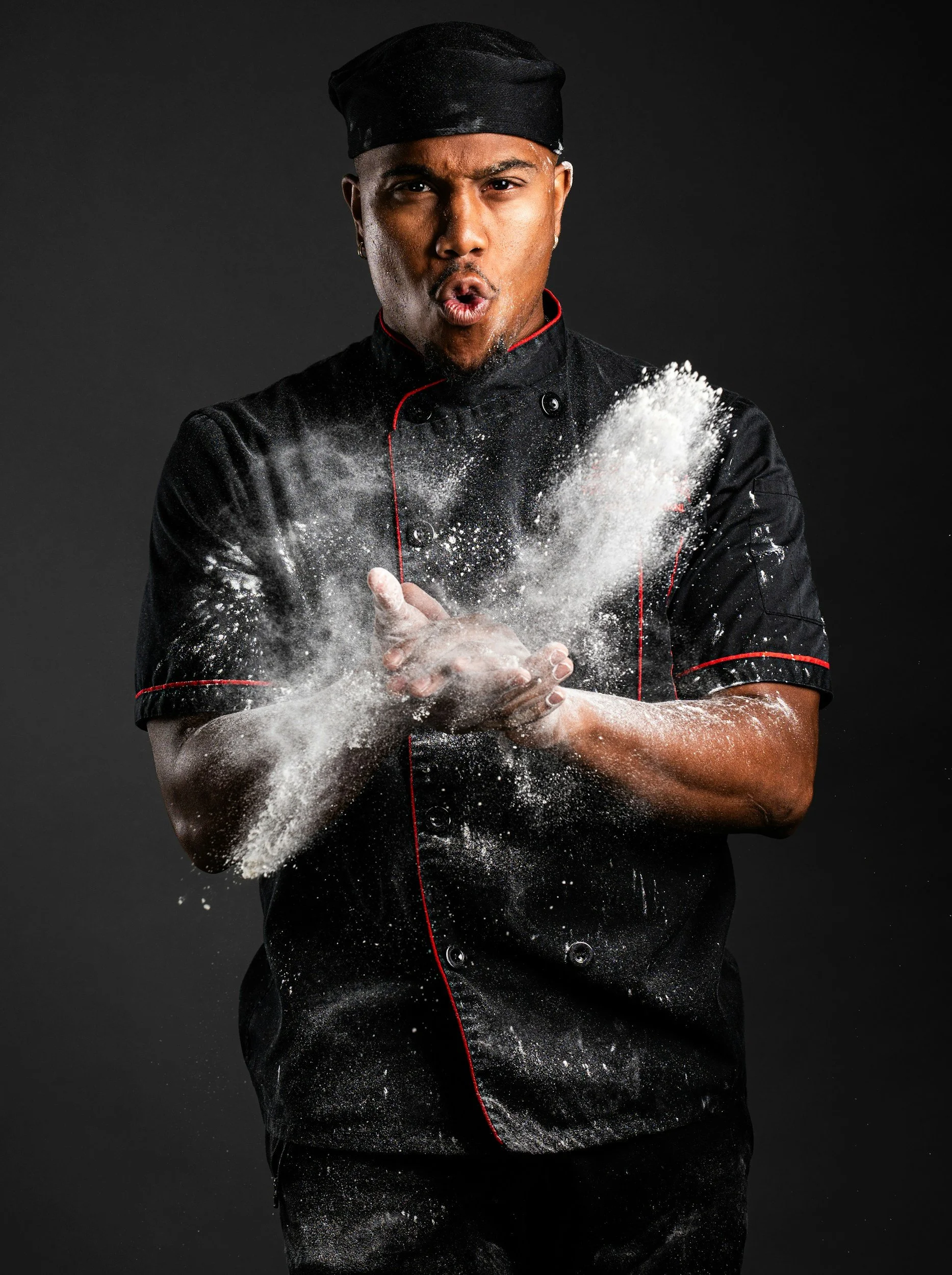 A man in a black chef's uniform with red piping, wearing a black chef's hat, is expressing surprise or shock as white flour or powder disperses around his chest and hands against a dark background.