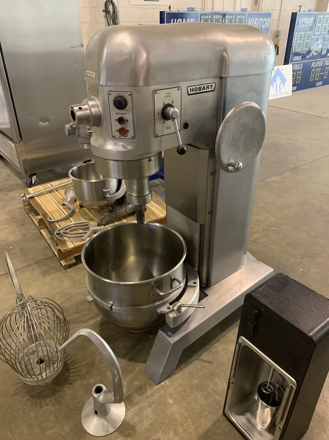 A commercial Hobart stand mixer with a large mixing bowl attached, surrounded by kitchen equipment and utensils in a warehouse or industrial setting.