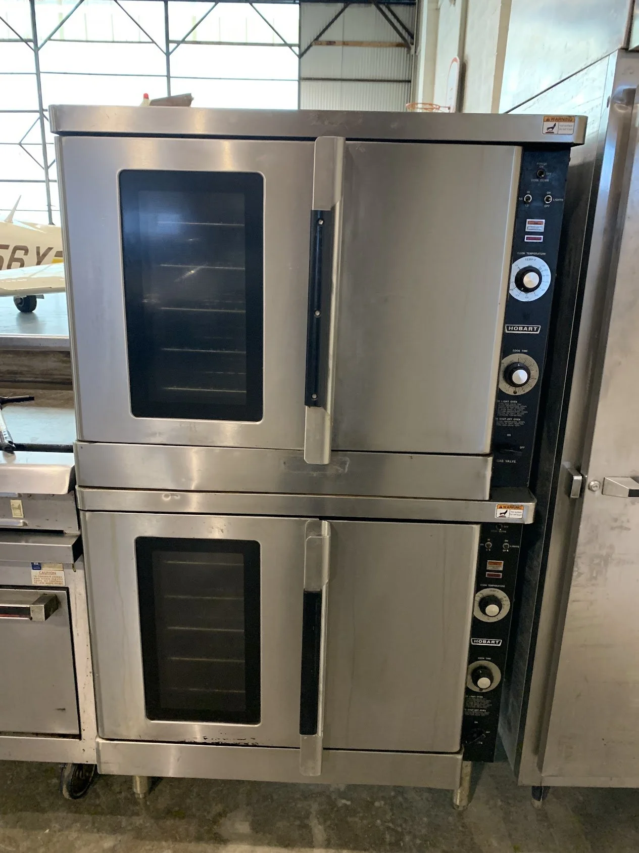 Two stainless steel commercial convection ovens, stacked one on top of the other, each with glass window doors and control knobs on the right side, located in a spacious industrial kitchen or warehouse with high ceilings and large windows.