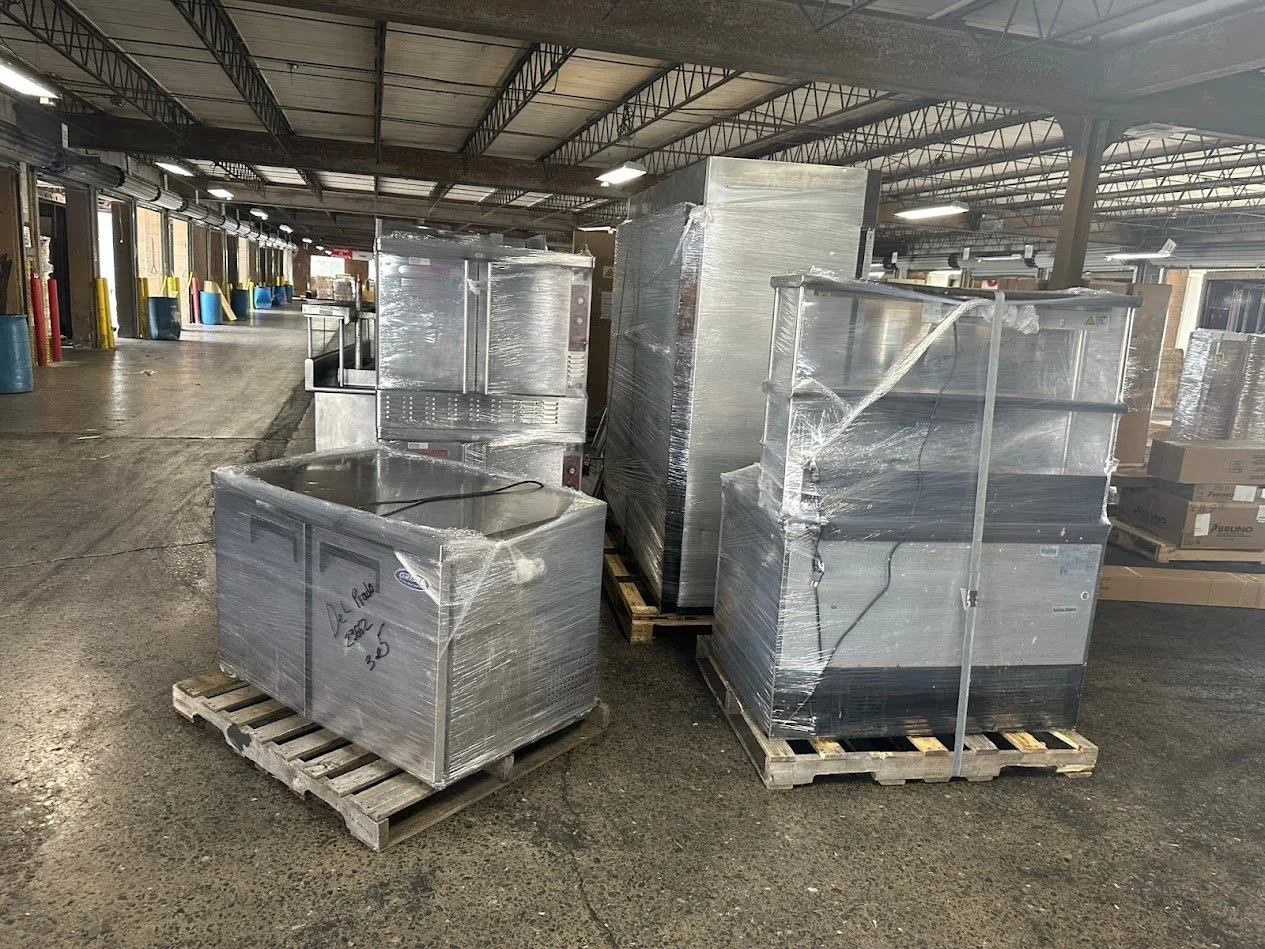 Warehouse with industrial kitchen equipment wrapped in plastic on pallets, concrete floor, metal ceiling, and yellow, red, blue safety cones along the wall.