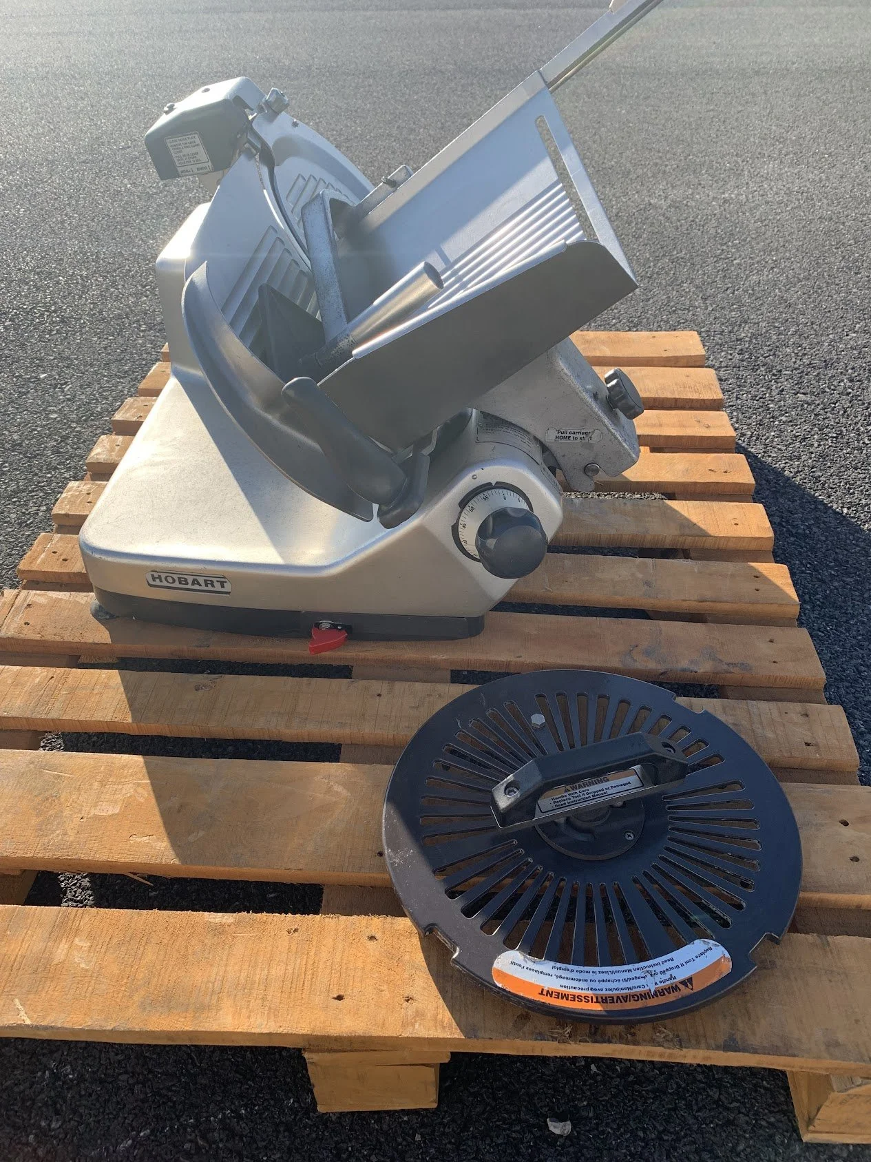 A commercial meat slicer on a wooden pallet outdoors with a detached blade guard in front of it.