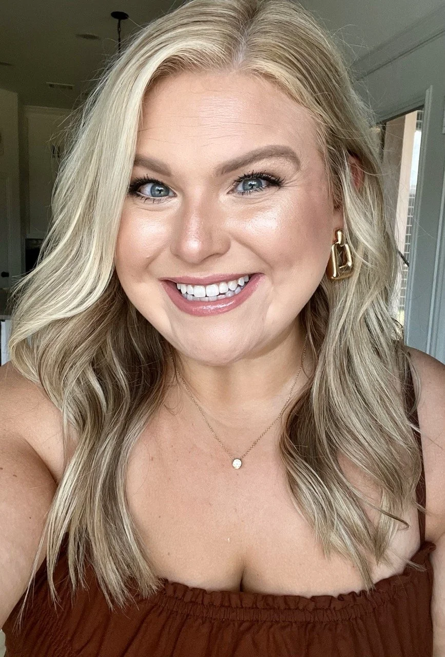 Close-up of a smiling woman with blonde wavy hair wearing gold earrings, a delicate necklace with a small pendant, and a brown top.