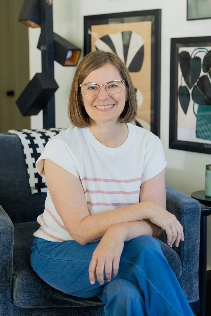 A woman with shoulder-length brown hair wearing glasses, a white t-shirt with pink stripes, and blue pants, sitting on a dark blue armchair, smiling in a modern living room with black and white artwork and a black lamp in the background.