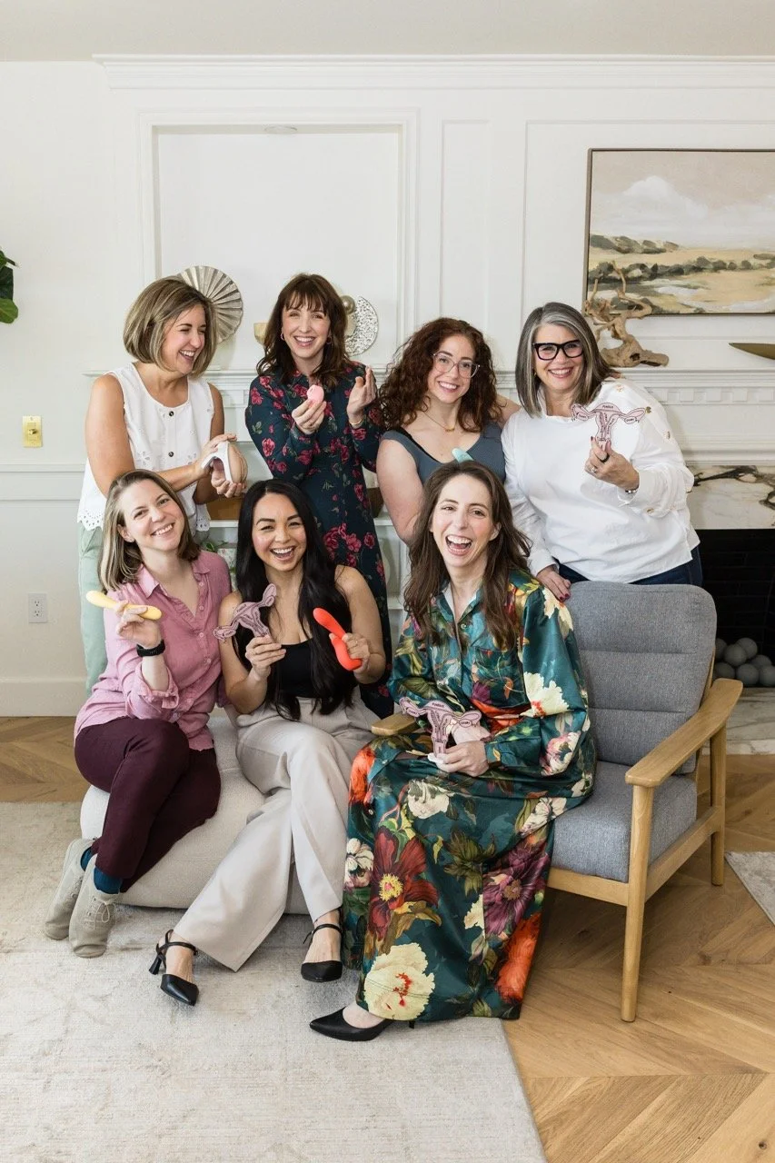 Group of seven women smiling and laughing in a living room, holding decorative animal-shaped objects, with a couch and artwork in the background.