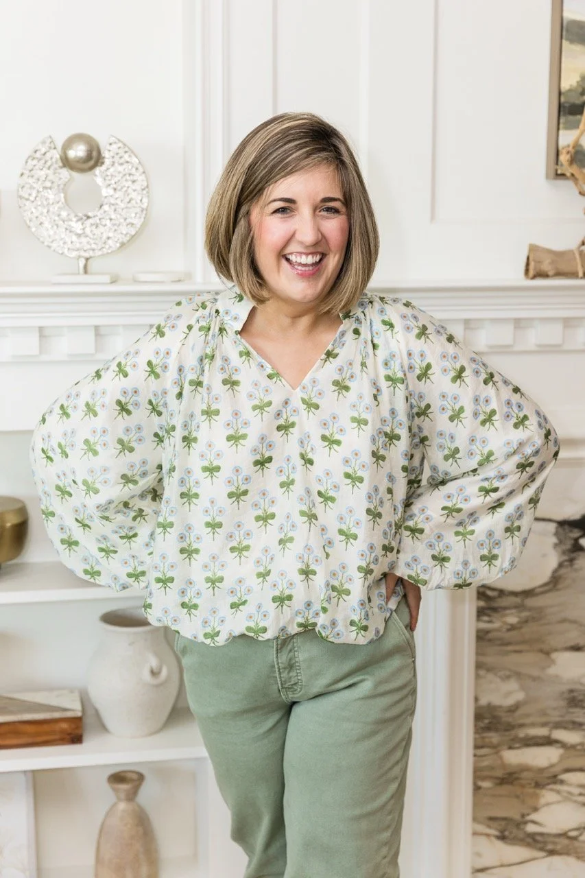 A woman with short blonde hair smiling, wearing a white top with green and light blue floral pattern and light green pants, standing in front of a white decorated wall.