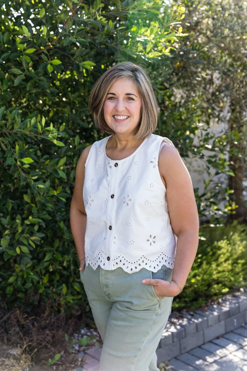 A woman standing outdoors in front of green bushes, smiling, wearing a sleeveless white eyelet blouse and light-colored pants.