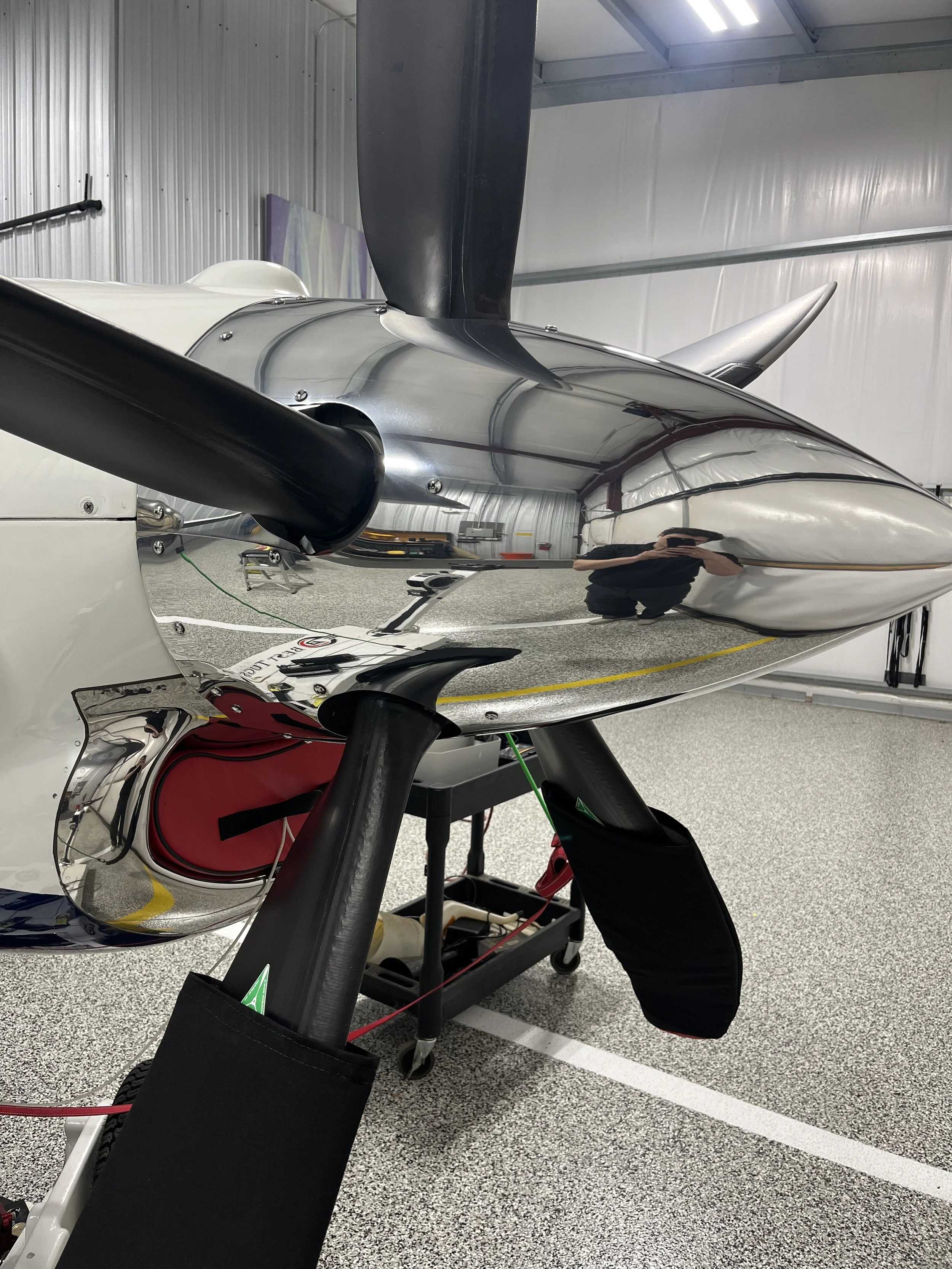 A close-up view of a sleek, futuristic racing aircraft with a polished, reflective surface inside a hangar. The aircraft features black and white colors with a sharp, aerodynamic design, including a prominent black wing and a pointed nose. The hangar