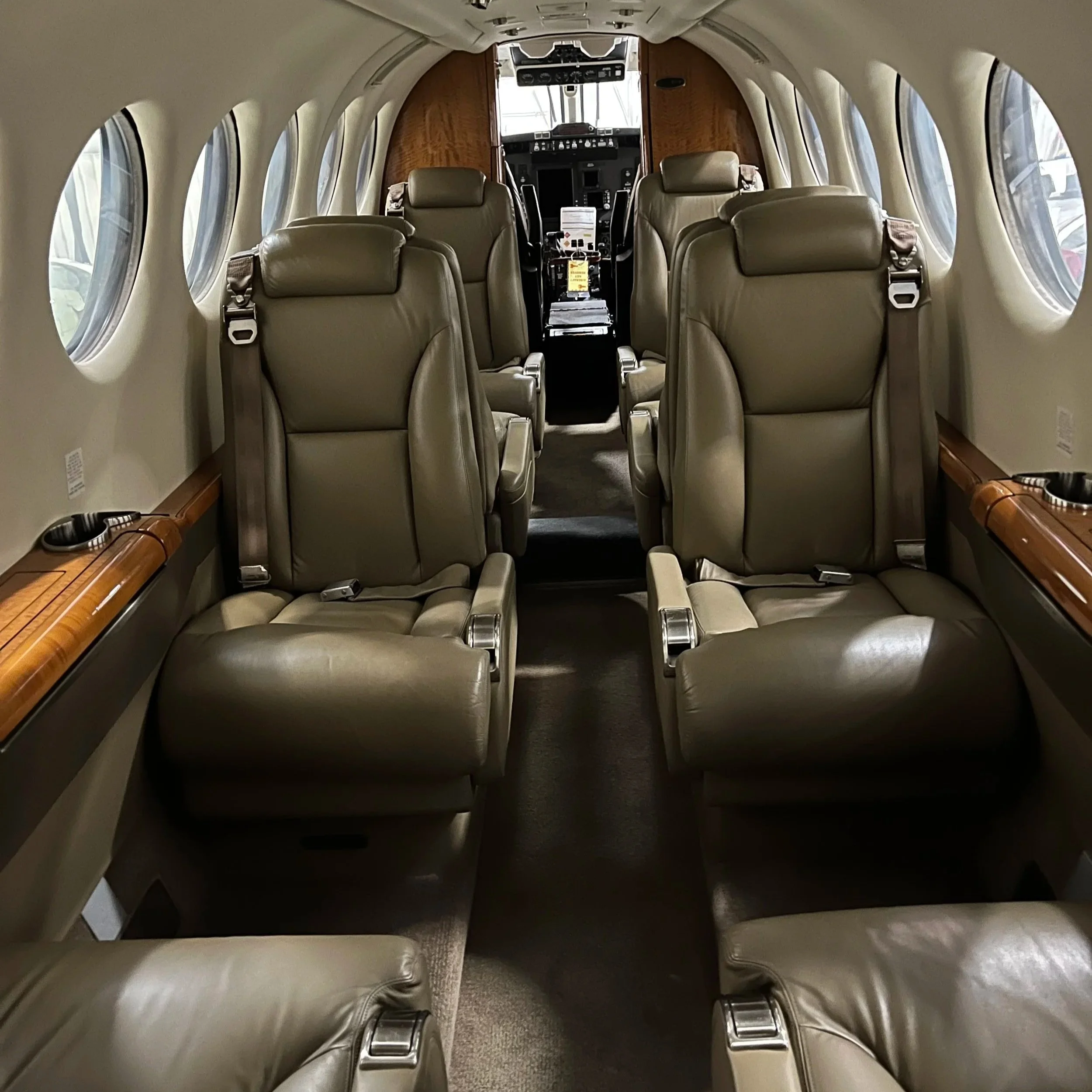 Inside view of a small private airplane cabin with beige leather seats, large oval windows lining both sides, and the cockpit visible in the front.