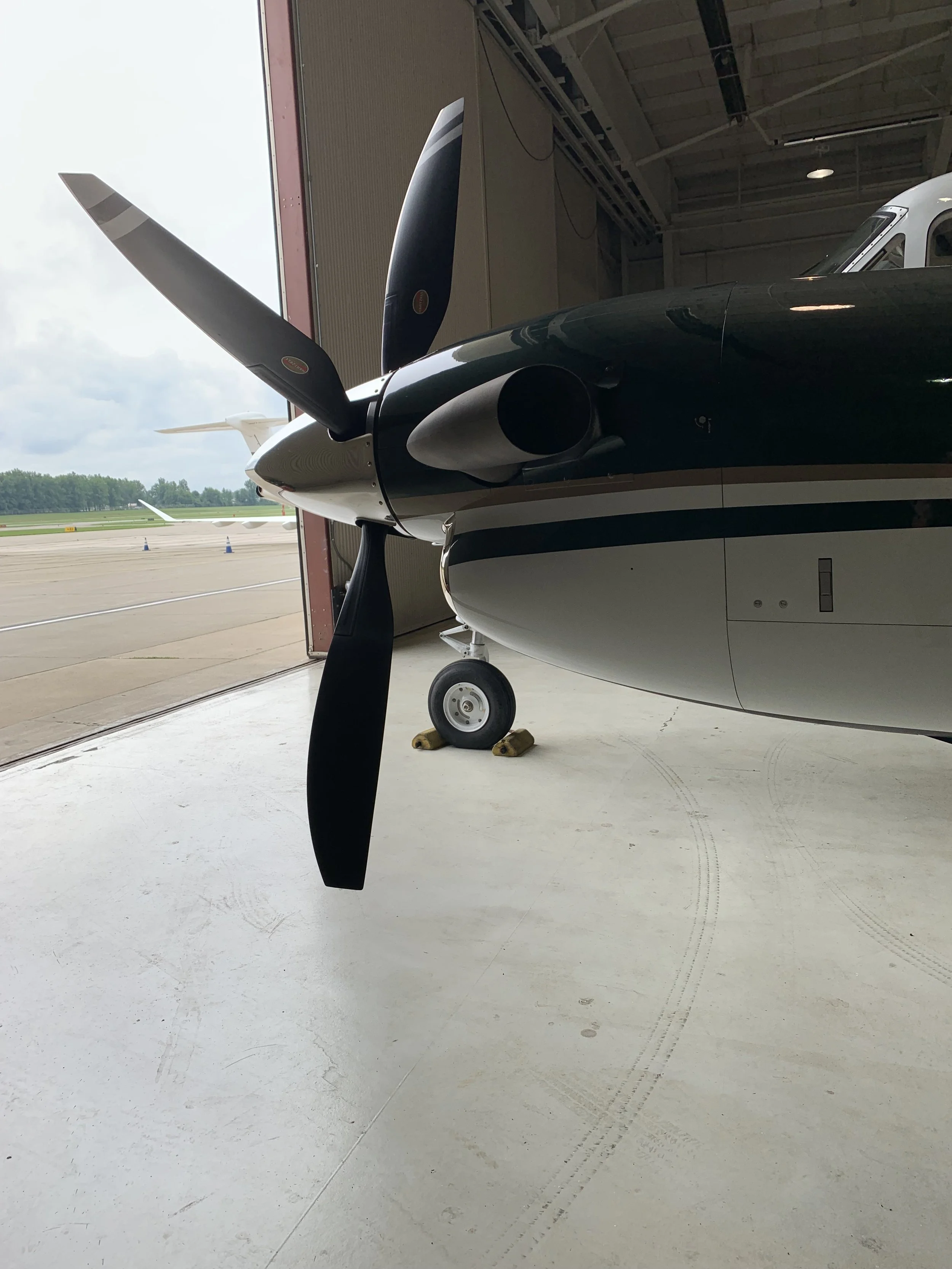 Small aircraft inside a hangar with its nose wheel on chocks, under the open hangar door showing a view of the runway and distant planes.