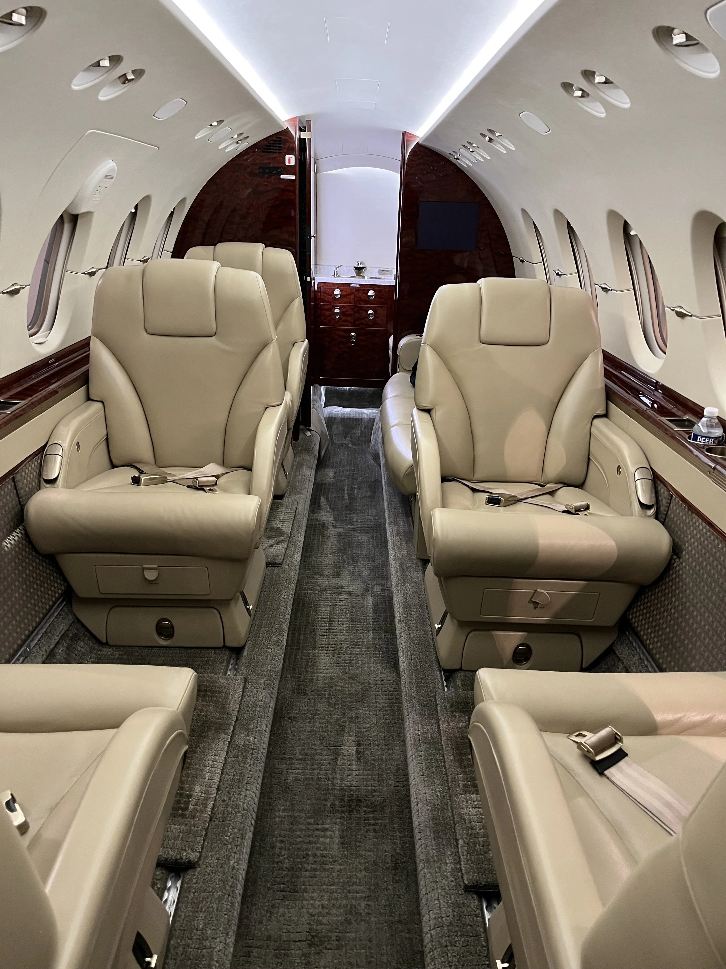 Interior of a private jet with beige leather seats, windows lined along the side, and wood paneling, seen from the back of the plane.