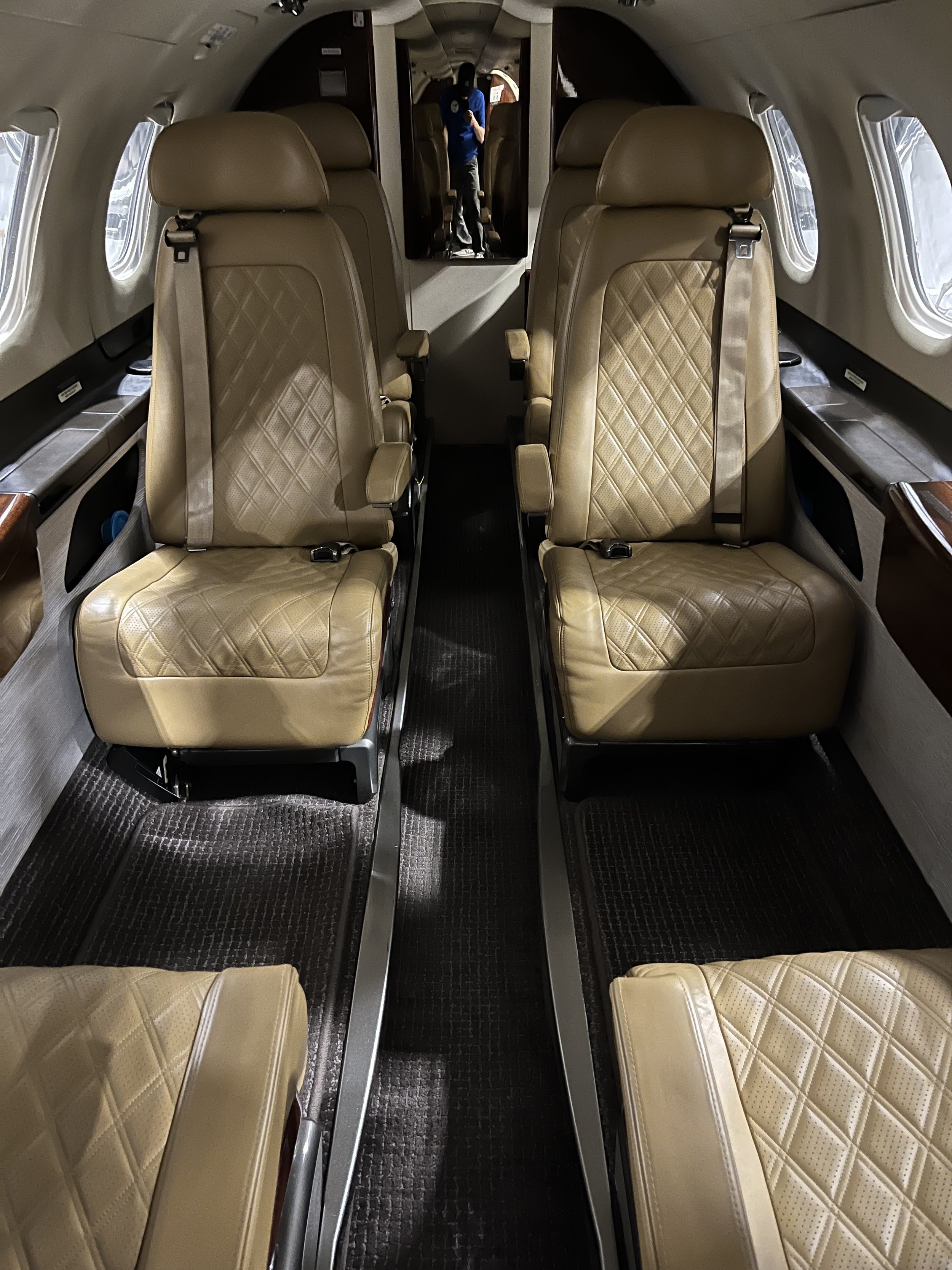 Interior of a small airplane with beige leather seats arranged in pairs along a narrow aisle. The seats have diamond-quilted stitching and headrests. Small windows line the aircraft walls, and there is a mirror on the back wall showing a person in a 