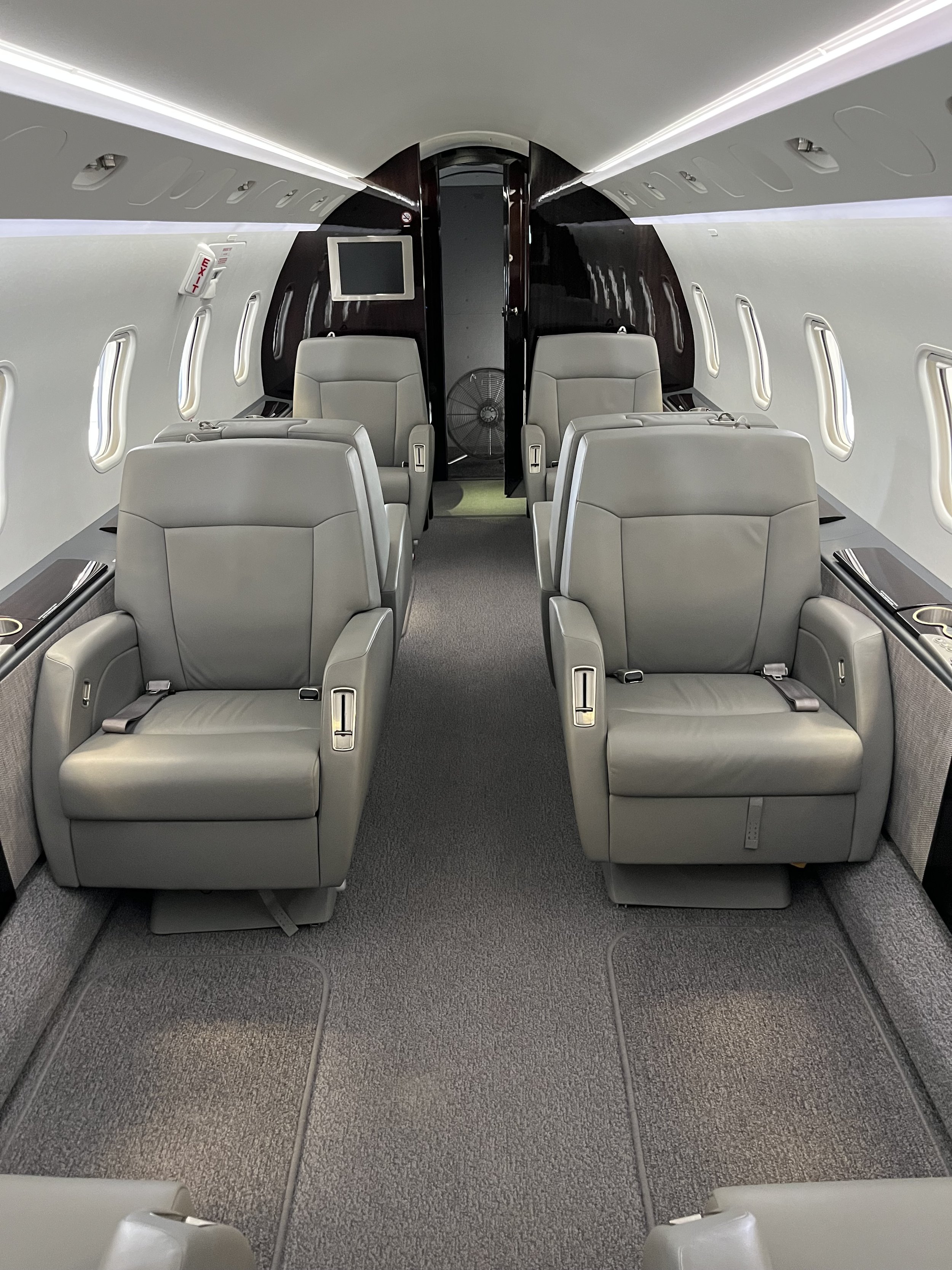 Interior of an airplane cabin with four gray leather seats arranged in two rows facing forward, a small display screen on the dark wood wall at the front, and multiple windows along the side walls.