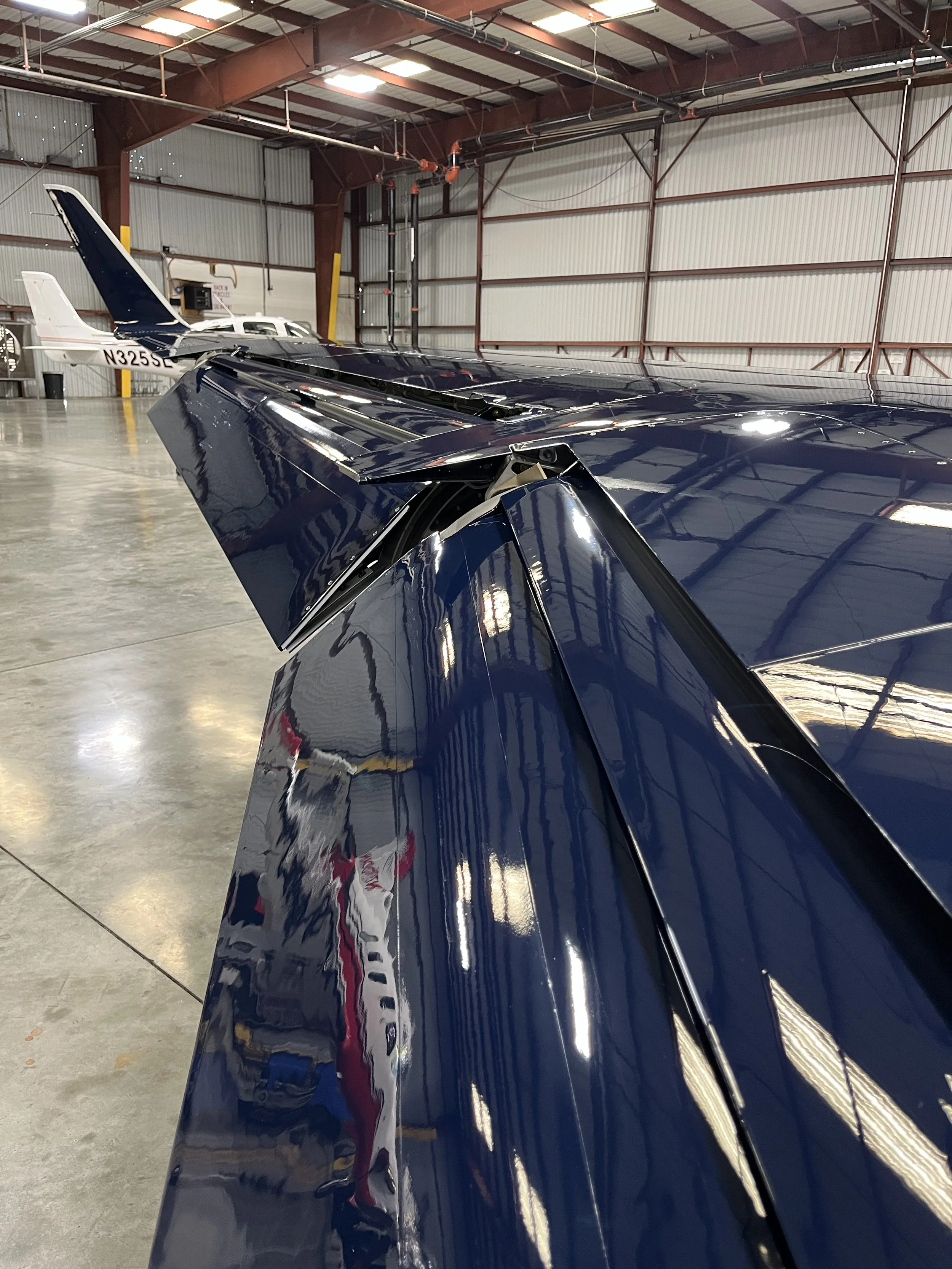 Close-up of a shiny, dark blue airplane wing with a reflection of a person wearing a red shirt visible on its surface, inside an airbase hangar with a corrugated metal ceiling and another small aircraft in the background.