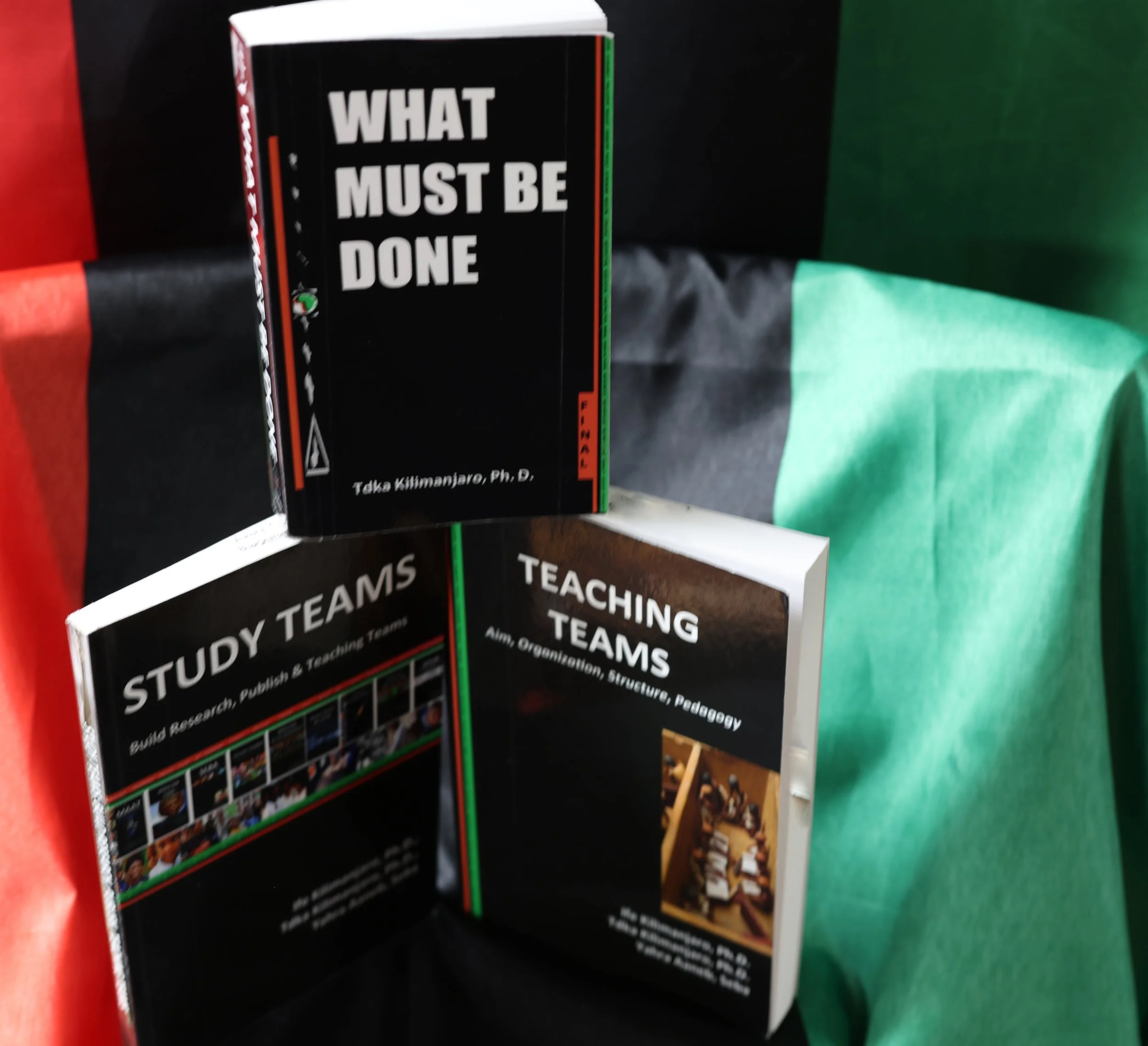 Close-up of a black book titled "What Must Be Done" with the author’s name, Taka Kilimanjaro, Ph. D., visible at the bottom of the cover. The book is standing upright on a surface, behind other books with colorful covers, against a background of black, green, and red fabric.