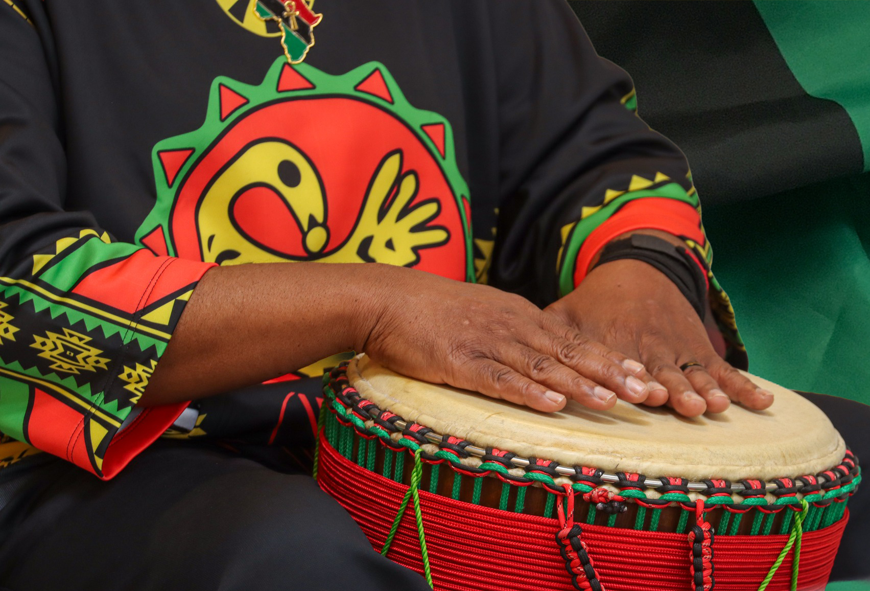 Person playing a drum with their hands, wearing a colorful shirt with a cartoon face and African pattern details.