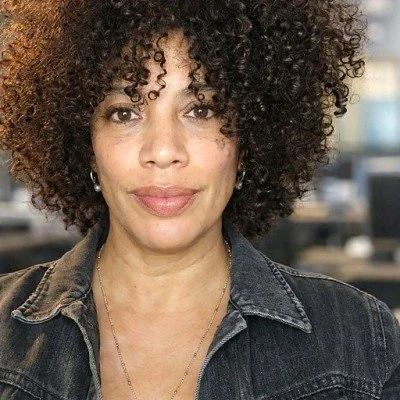 A woman with curly hair wearing a denim jacket and small earrings, standing indoors.