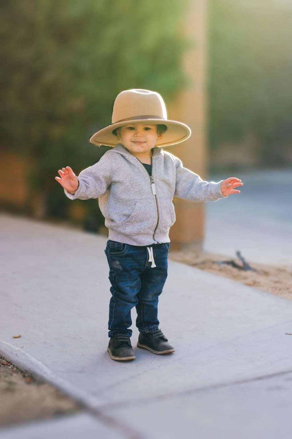 A young child standing on a sidewalk wearing a large beige hat, gray zip-up hoodie, dark jeans, and black shoes, with both arms slightly raised.