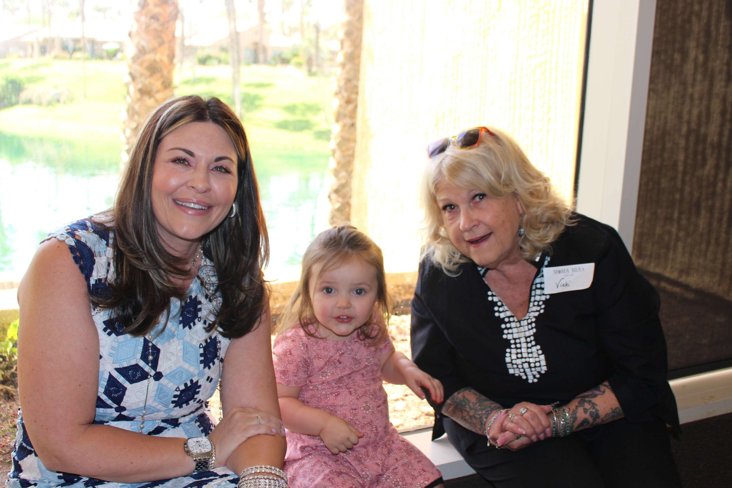 A group of four females and one young girl smiling and posing for a photo indoors, with large windows showing a grassy outdoor scene in the background.
