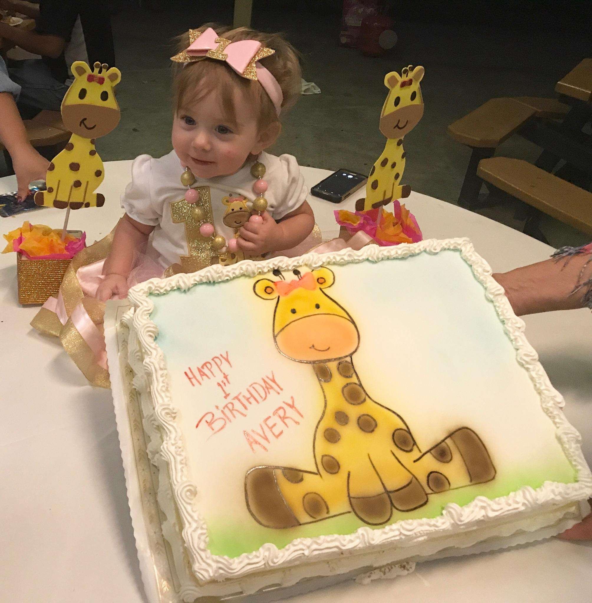 A young girl with curly red hair wearing a pink and gold bow headband, a white dress, and a necklace, is sitting in front of a birthday cake decorated with a giraffe illustration. The cake has a white icing border and a pastel green background. Two yellow giraffe-shaped decorations with smiling faces are on either side of the girl, with pink and yellow tissue paper accents around the bases. The girl is smiling and holding a small giraffe-shaped cookie or decoration. The setting appears to be a birthday celebration with a dark background and a table with gift bags or presents.