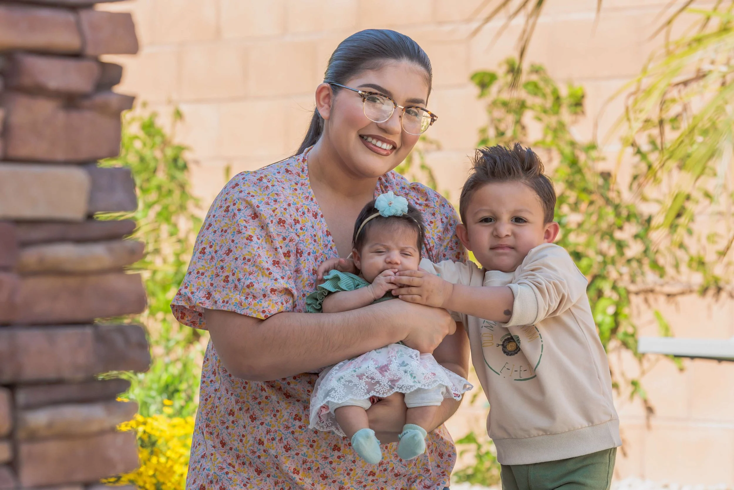 A woman with glasses holding two young children outdoors, smiling. One child has a flower headband, and the other is a boy with a serious expression. They are standing near a brick wall with green plants and yellow flowers in the background.