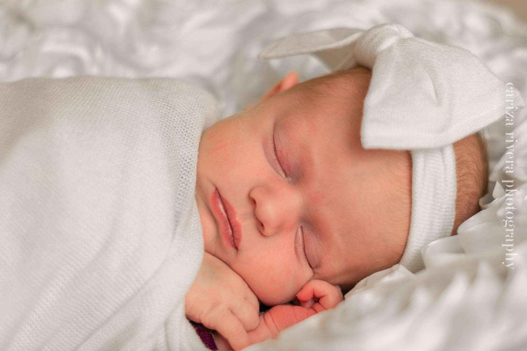 Close-up of a sleeping newborn baby with a white blanket and hat, resting peacefully with hands near the face.