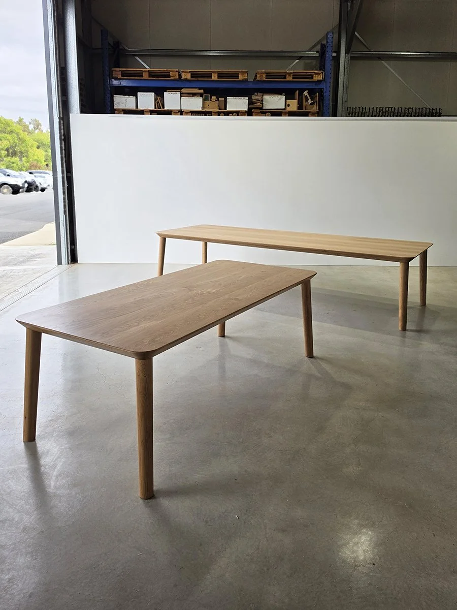 Two Mercator Way Tables in American Oak side by side in the workshop with roller door open to greenery outside - bespoke furniture by Nathan Day Design